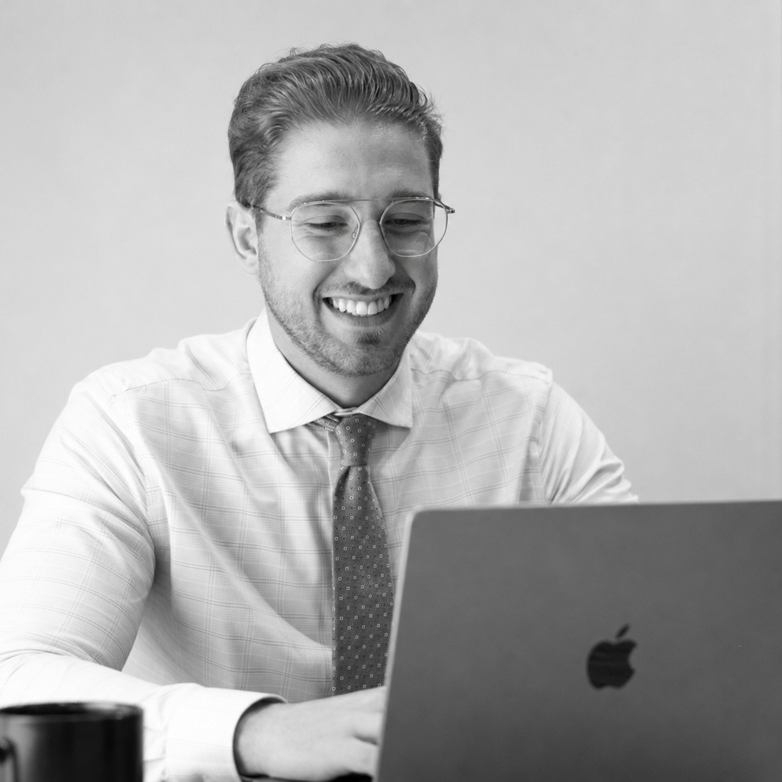 Jacob Suski smiling with glasses working on a laptop, wearing a dress shirt and tie.